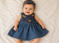 Fröhliches Baby trägt ein blaues Guess Jeanskleid mit farbenfroher Blumenstickerei und Schleife auf hellem Hintergrund. Stylisches Babykleid in Größe 62/68 mit verspieltem Denim-Look.
