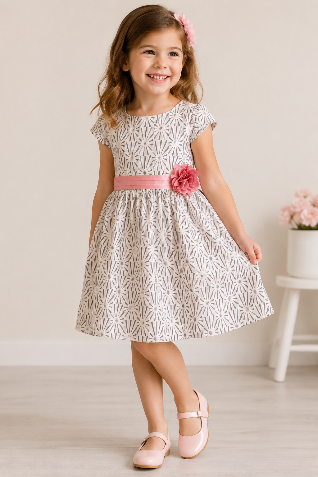 Lächelndes Mädchen trägt ein elegantes festliches Kleid mit modernem Schwarz-Weiß-Muster, roséfarbenem Satinband und Blütenapplikation. Stilvolles Sommerkleid für Kinder in Größe 128.