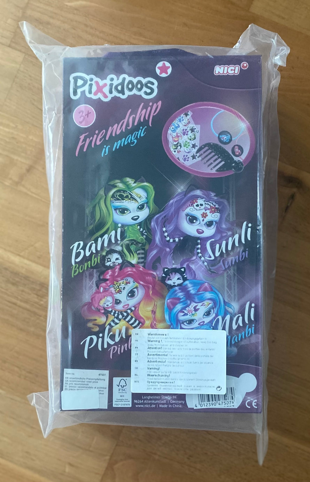NICI Pixidoos Piku Plüschtier in Originalverpackung auf weißem Hintergrund. Bunte Sammelfigur mit großen Augen, neonfarbenen Haaren und 3-teiligem Zubehörset. Ideal für Kinder und Sammler. Rückseite
