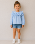 Mädchen trägt eine hellblaue Polo Ralph Lauren Bluse mit Rüschen und langen Ärmeln in Größe 104. Neuwertige Kinderbluse in elegantem Stil kombiniert mit Jeansshorts.