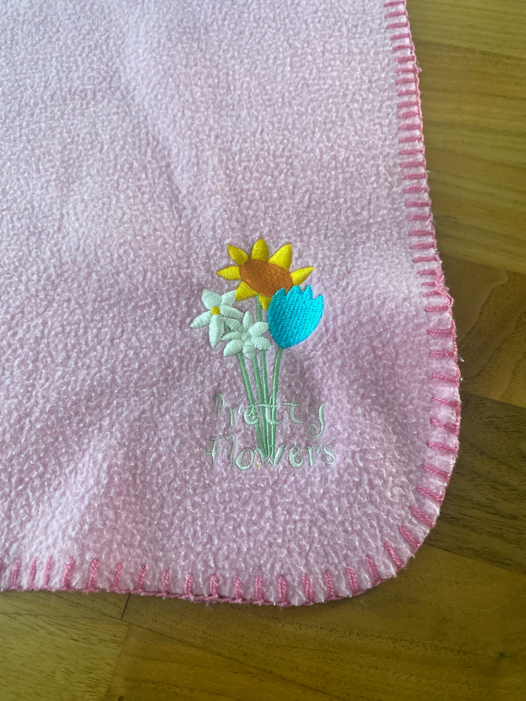Rosa Babydecke aus weichem Fleece mit dekorativer Blumenstickerei in der Ecke. Kuschelige Kinderdecke mit eingefasstem Rand, ideal für Kinderwagen, Bettchen oder gemütliche Kuschelstunden. Detailansicht