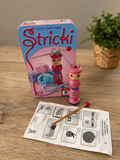 Stricki Strickspiel von Schmidt mit Metallbox, zwei Holz-Strickfiguren, Stricknadel und Original-Anleitung auf Holztisch.